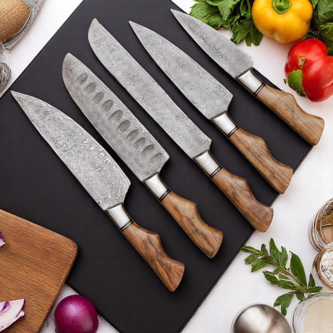 Five handmade Damascus steel chef knives with light wood handles and wavy patterns, arranged on a black cutting board with fresh vegetables