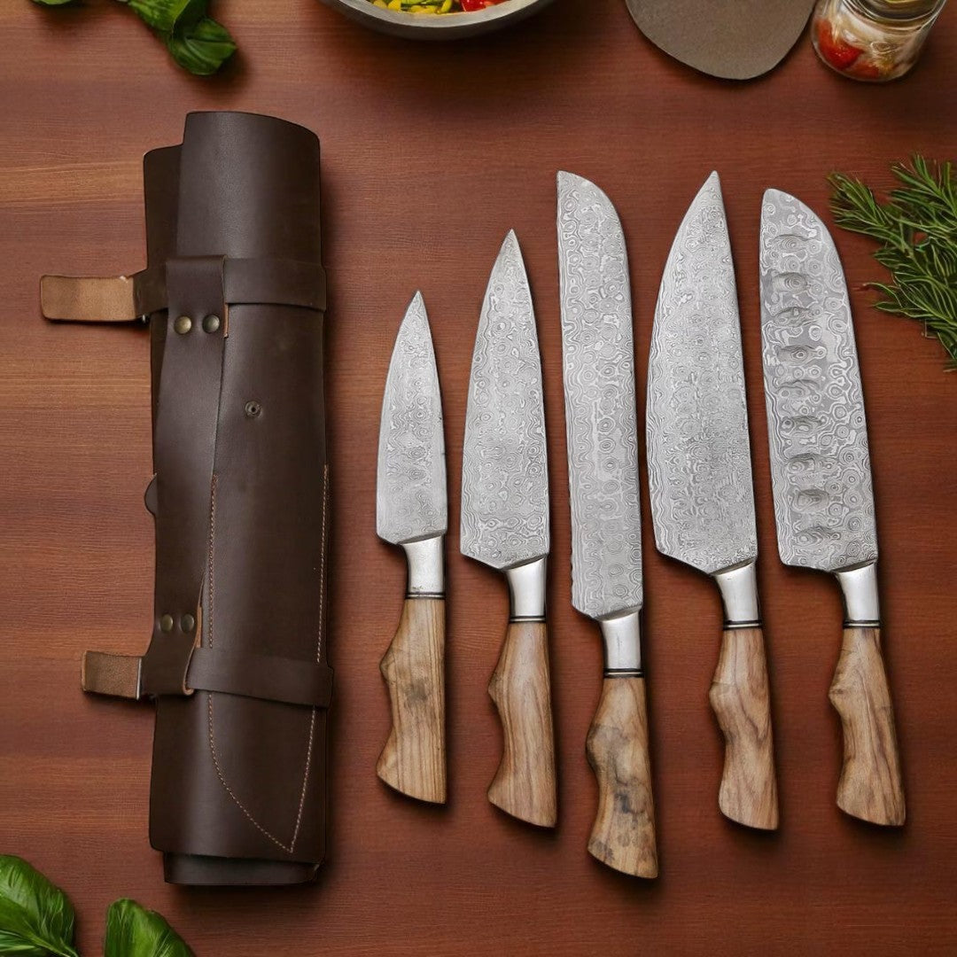 Overhead view of five handmade Damascus steel chef knives and their brown leather roll bag, arranged on a wooden table with kitchen ingredients