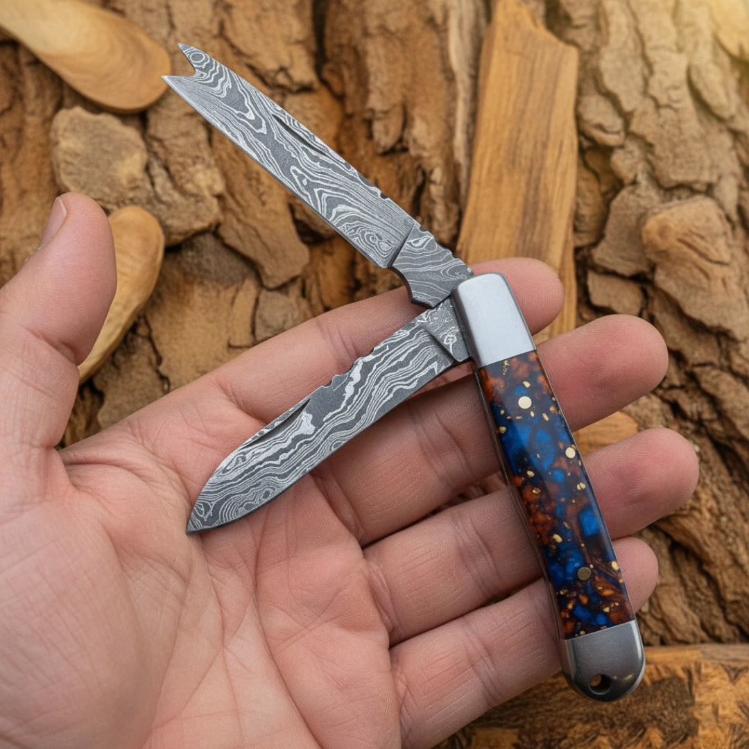 A hand holding a 7.5-inch handmade Damascus folding bull cutter knife with the blades open, displaying the detailed Damascus steel patterns and the blue resin handle design.
