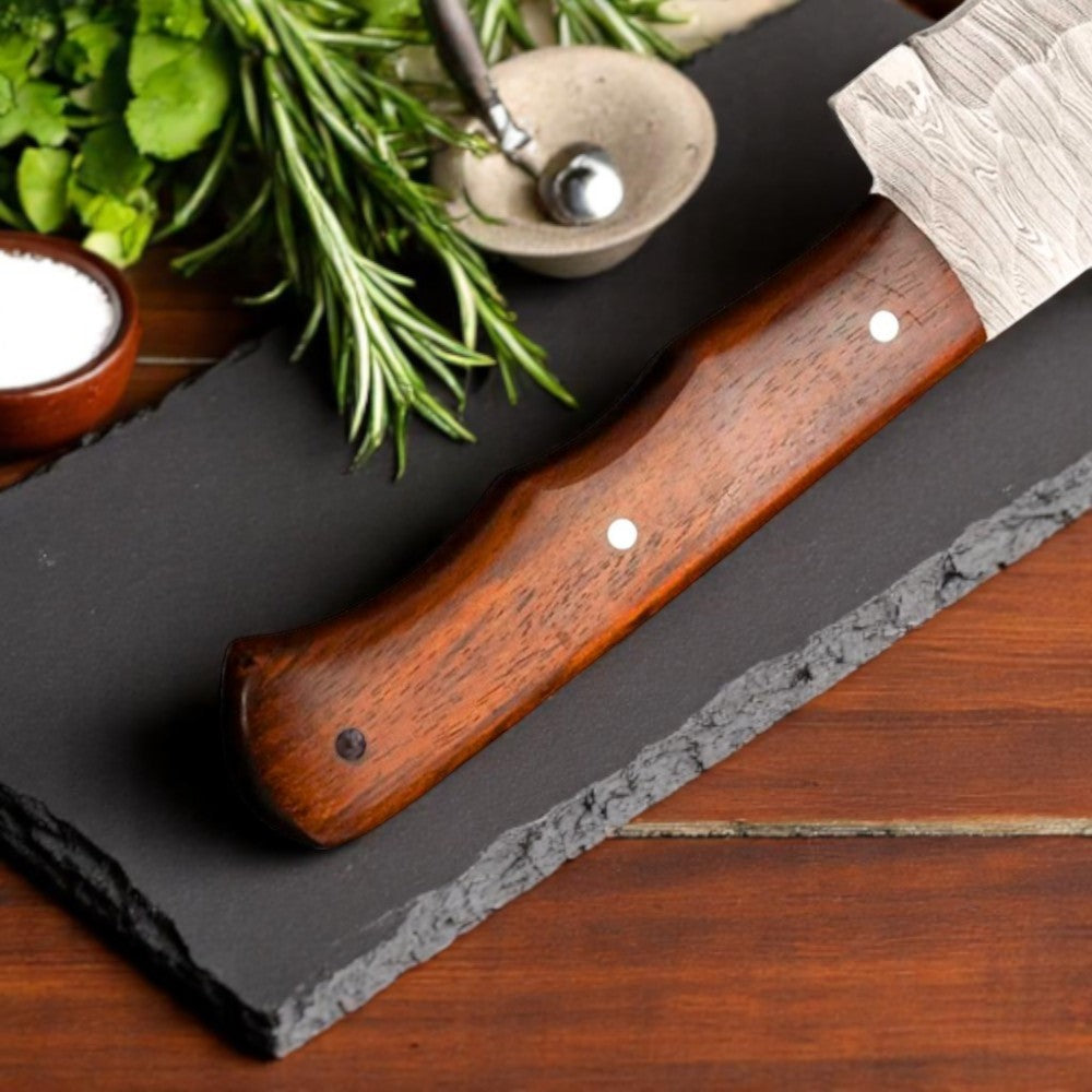 Bold Clyvano 12-inch Damascus Brisket Knife with Rose Wood handle partially in its brown leather sheath, on a black slate board with rosemary

