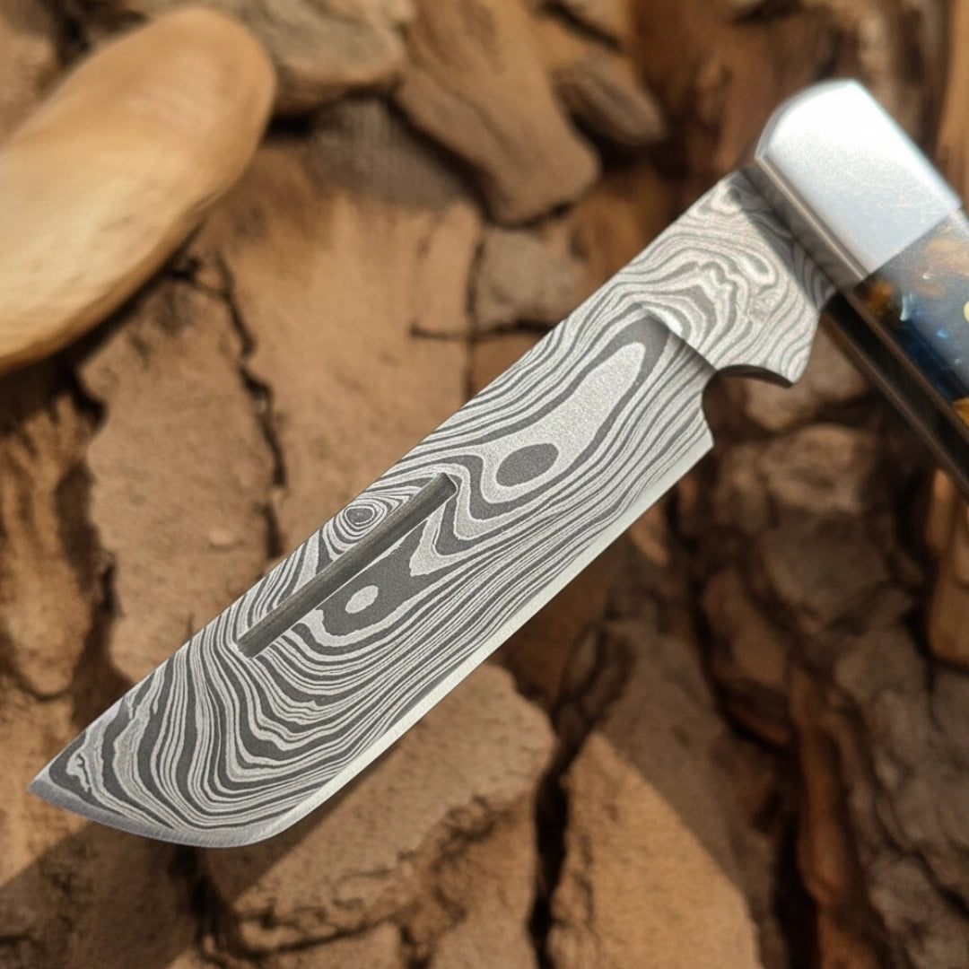 A detailed view of the Damascus steel blade of the Cowboy Bull Cutter folding knife, showcasing the unique wavy pattern of the steel, with the blue resin handle visible in the background.