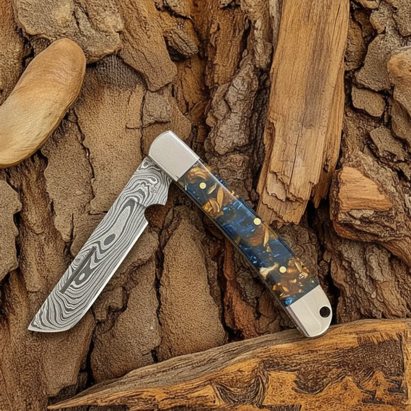 A close-up of a Cowboy Bull Cutter folding knife with a Damascus steel blade, highlighting its intricate pattern, resting on a textured leather sheath with a dark wood background.