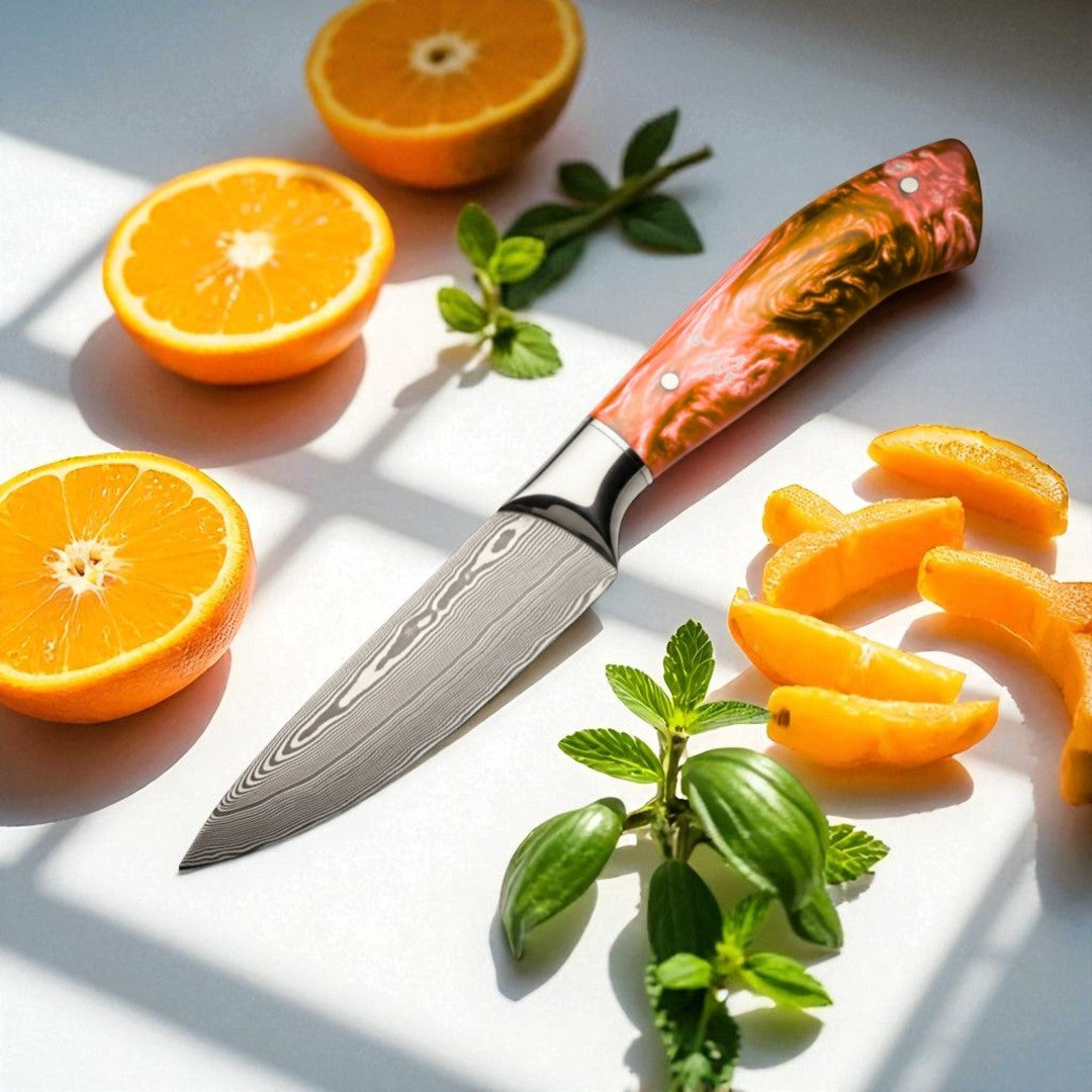 Damascus paring knife with an orange and brown swirled resin handle resting on a white surface with cut oranges and mint leaves.