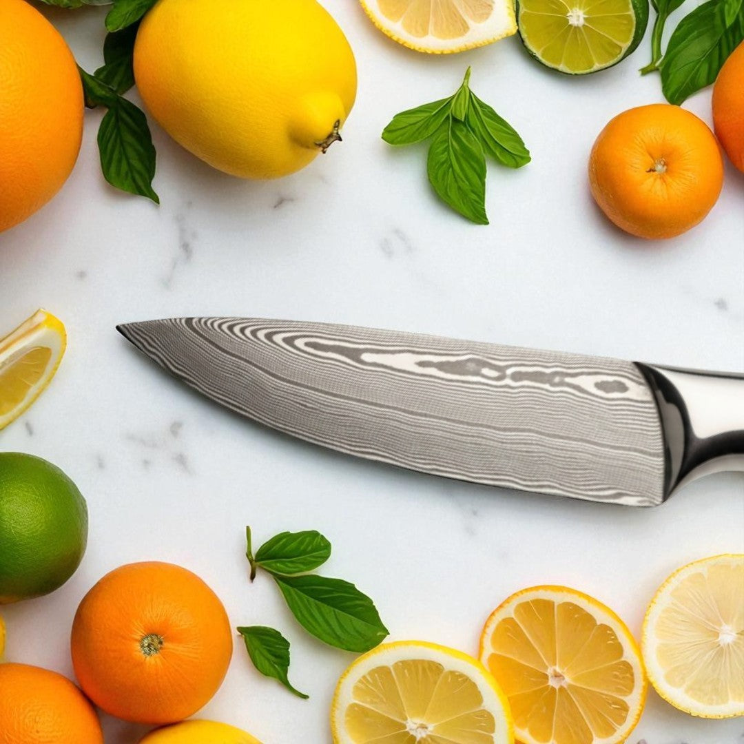 Close-up of the Damascus steel blade of a paring knife, surrounded by oranges, lemons, and limes on a white marble surface.