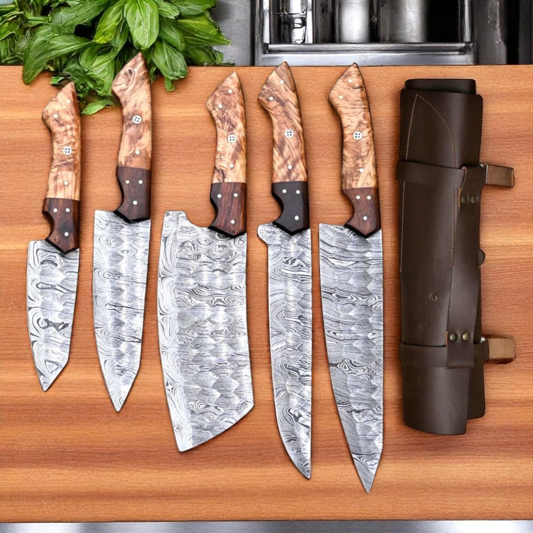 Five custom hand-forged Damascus steel chef knives with unique wood handles and twist patterns, displayed upright on a wooden cutting board next to a brown leather knife roll
