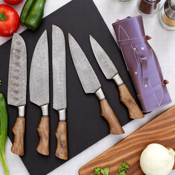Set of five Damascus steel kitchen knives with wooden handles on a black cutting board, accompanied by a purple knife roll and vegetables.