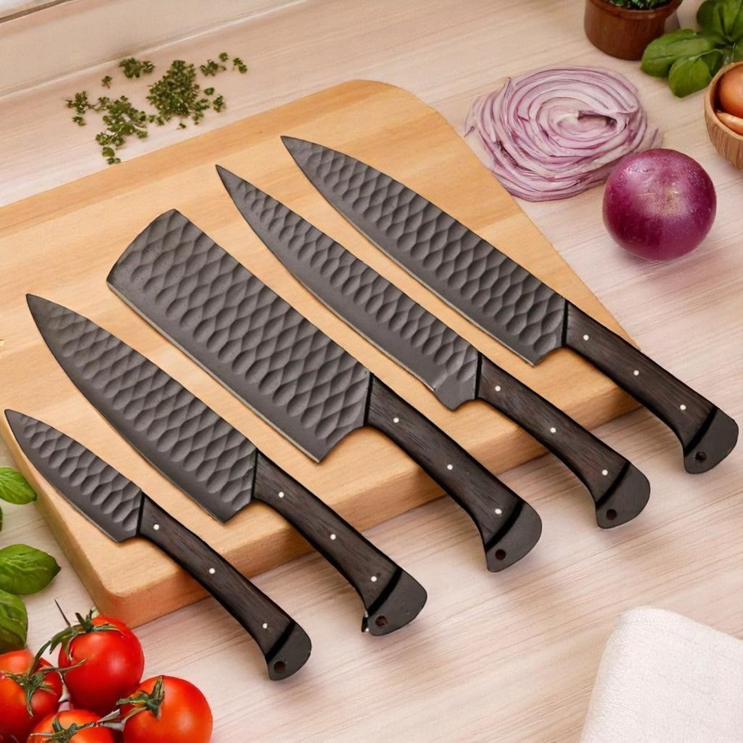 Five black hammered kitchen knives with dark wood handles and textured blades, fanned out on a light wooden cutting board with chopped herbs and red onion

