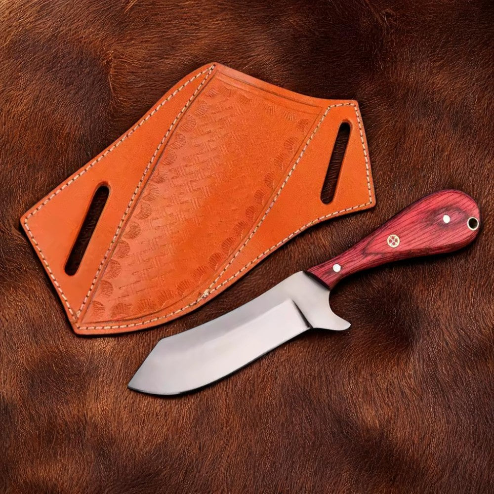 Handmade Carbon Steel Bull Cutter Knife with red Pakkawood handle, resting in its brown leather sheath on a wooden surface, showing decorative stamping.