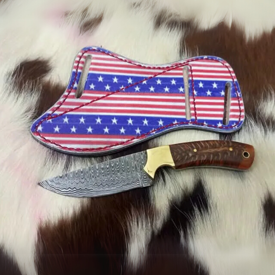 Damascus Cowboy Knife with wood handle and wavy blade, resting next to its USA Flag-themed leather sheath on a brown and white cowhide.