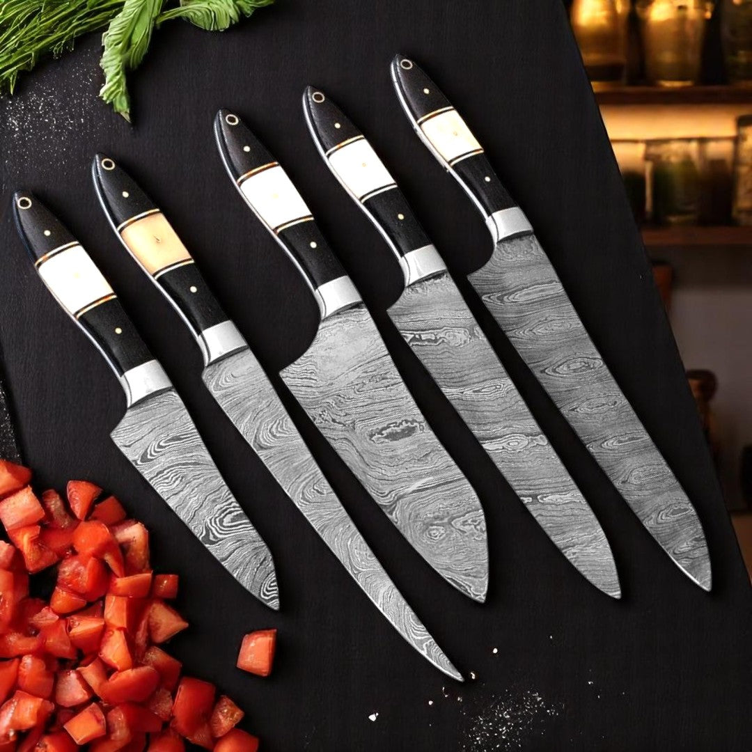 Overhead view of five Damascus chef knives with black wood and camel bone handles, arranged on a black cutting board surrounded by fresh vegetables