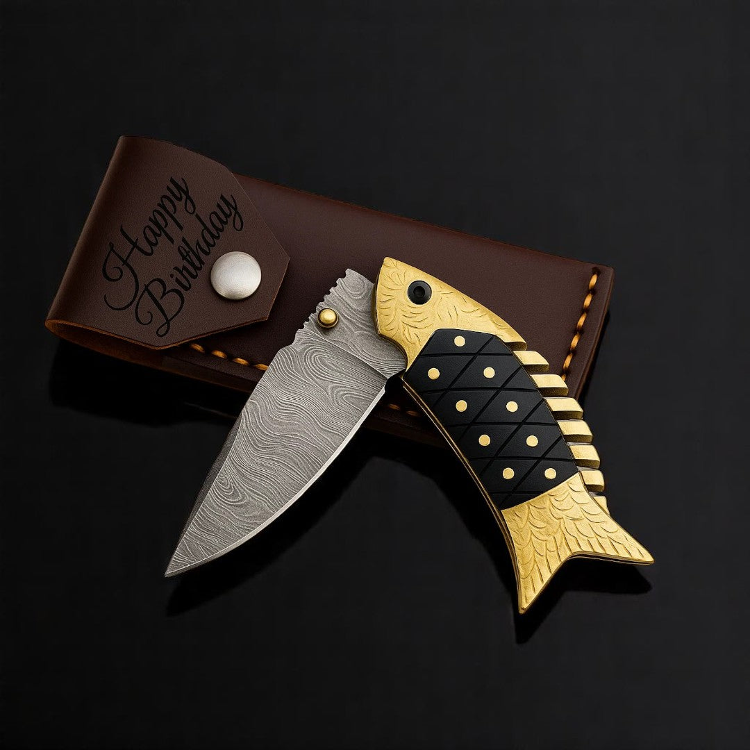 Close-up view of the gold-toned fish-shaped handle of a Damascus Folding Knife, showing black textured inlays and decorative gold dots.

