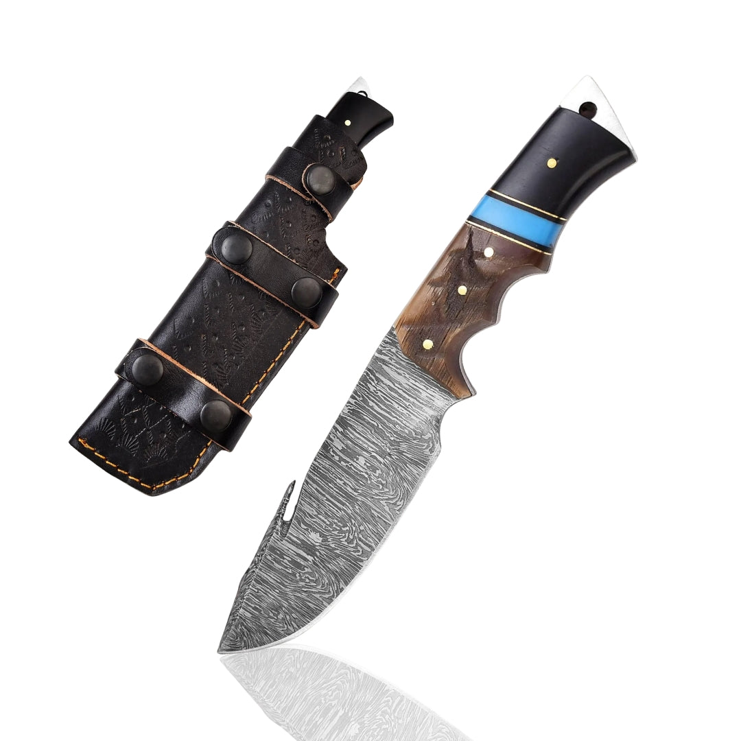 Damascus Gut Hook Skinner Knife with unique blue, black, and wood handle, resting on a wooden surface with pine needles and a brown leather sheath.