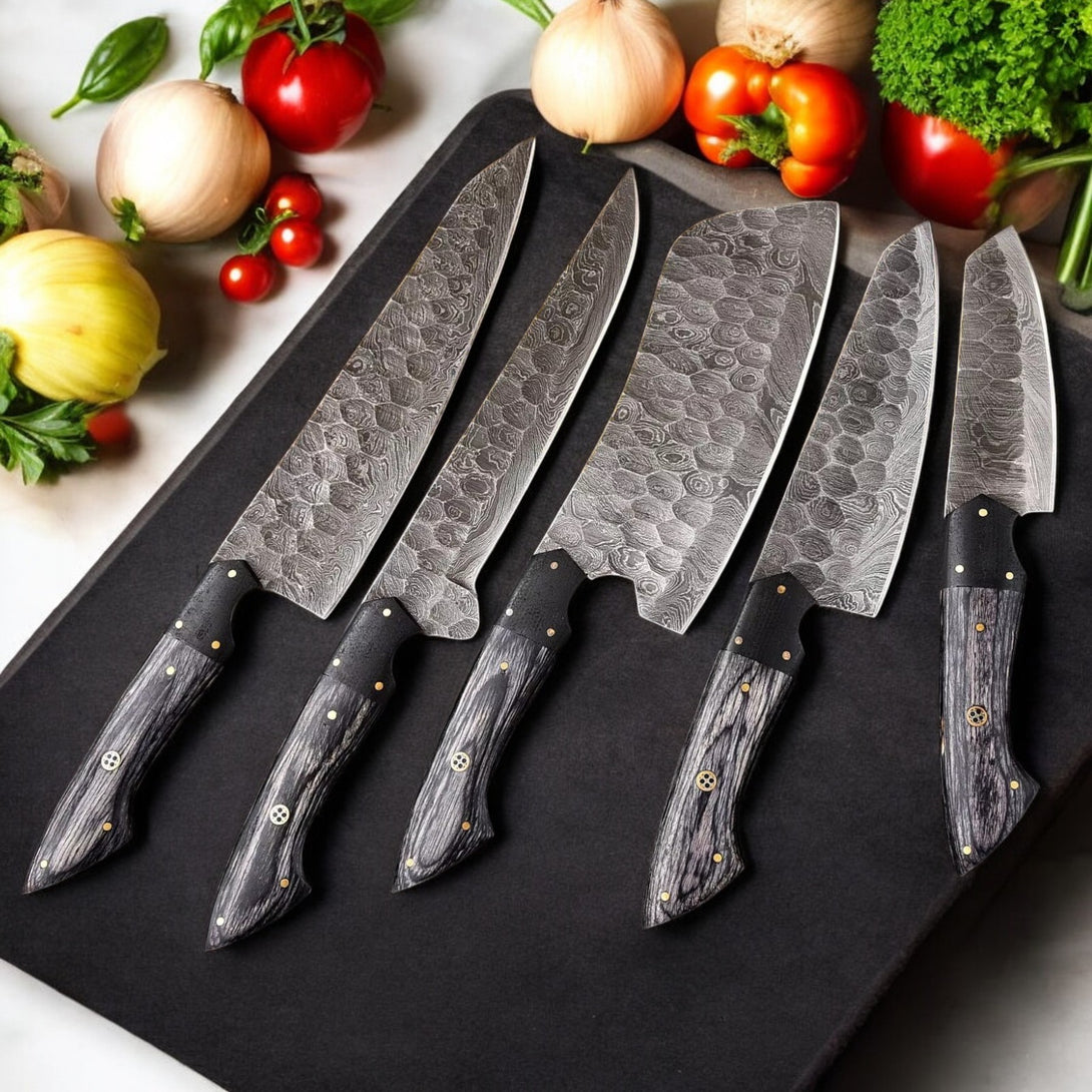 Overhead view of five Damascus steel kitchen knives with dark wood handles and brass accents, arranged on a black cutting board with fresh vegetables

