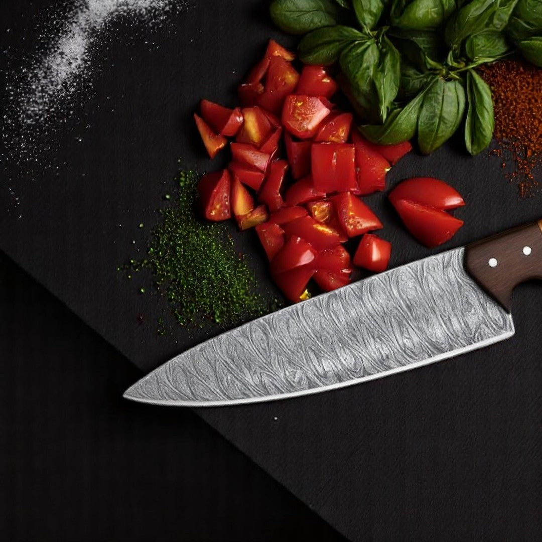 Japanese Damascus Chef Knife with wavy brocade blade and wood/resin handle, on a dark wooden surface with sliced tomatoes, cucumbers, and fresh herbs