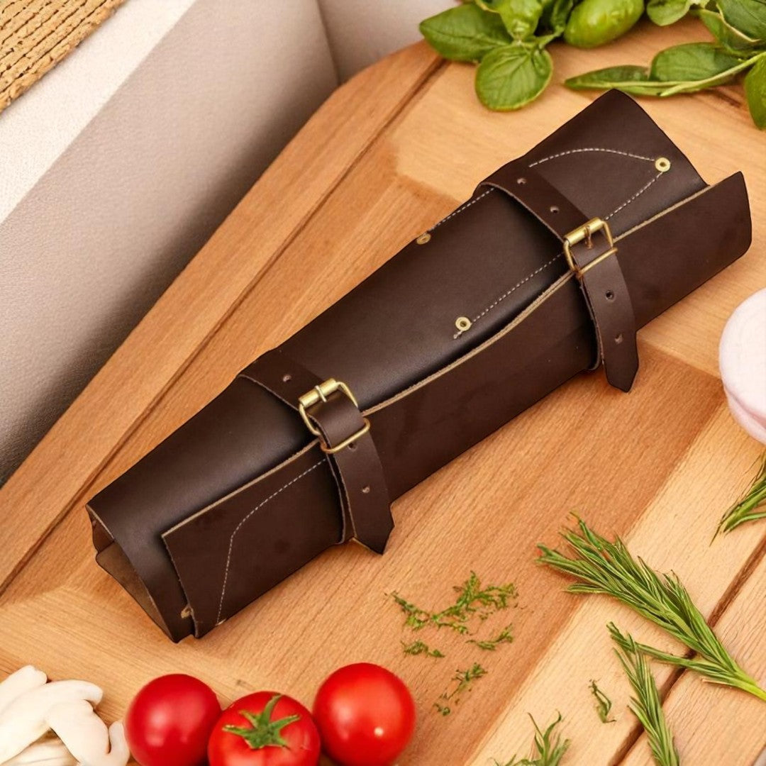 Five black hammered kitchen knives with dark wood handles and white rivets, displayed with their brown leather roll bag on a wooden cutting board with tomatoes and garlic

